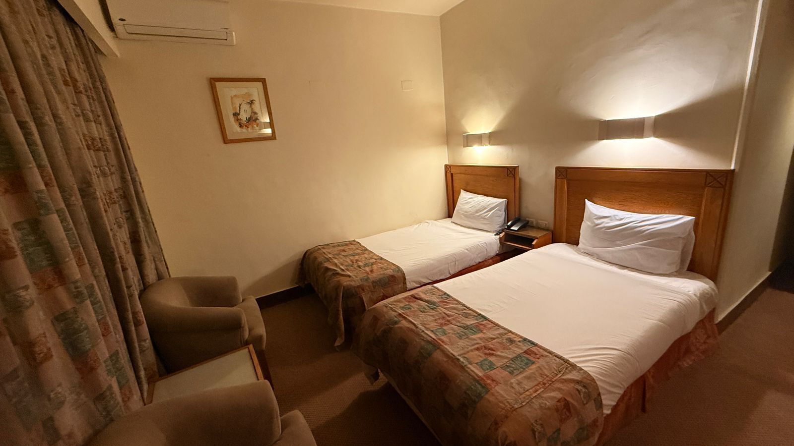 A hotel room with two beds, a chair, a nightstand, a lamp, and a painting on the wall.