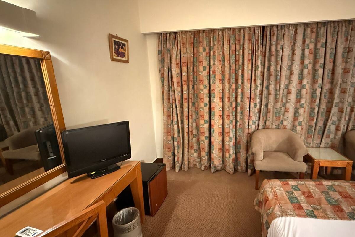 A hotel room with a bed, two armchairs, a small table, a television, a mirror, and a large window with curtains.