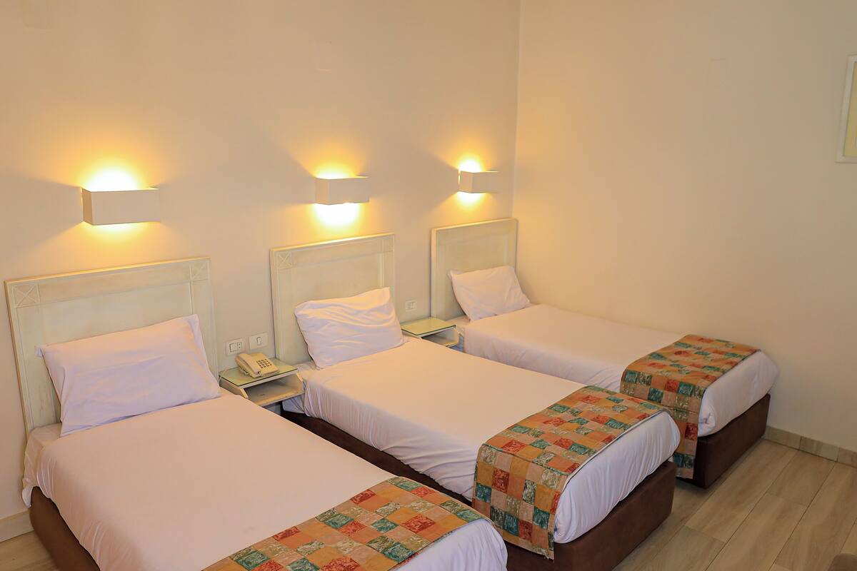 A hotel room with three beds, each with a white bedspread and a quilted blanket. There are bedside tables with lamps and a phone on the first bed.