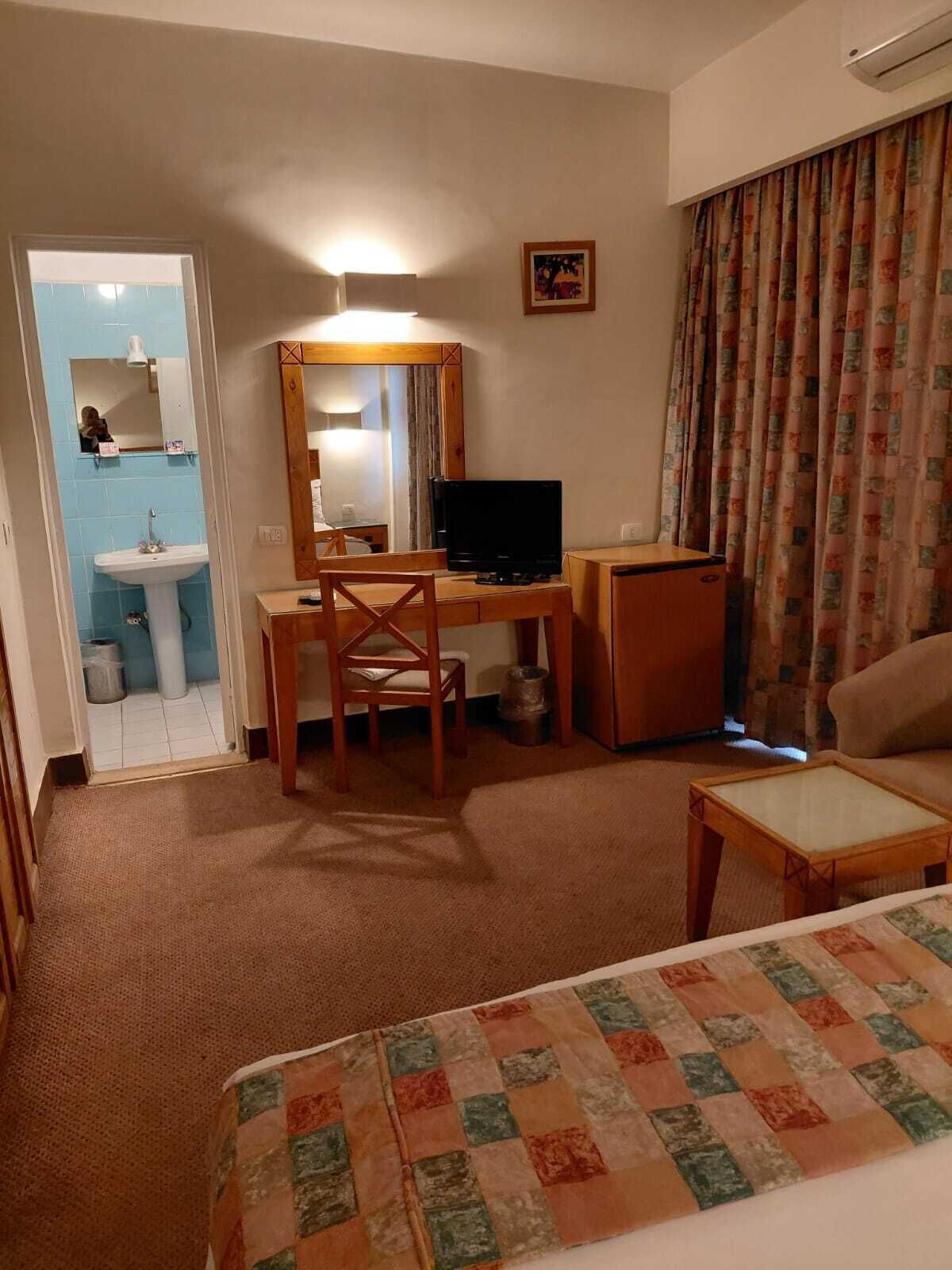 A hotel room with a bed, a desk with a chair, a television, a small table, a sofa, a bathroom with a sink, and a mirror.