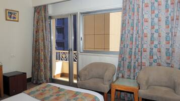 A hotel room with a bed, two armchairs, a small table, and a sliding glass door leading to a balcony.