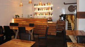 Un restaurante con mesas y sillas de madera, una zona de bar con estantes llenos de botellas, y lámparas colgantes.