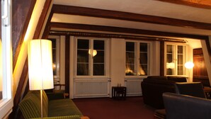 Un salón acogedor con un sofá verde, un sillón negro y una lámpara. Hay dos ventanas con marcos blancos y un radiador entre ellas.