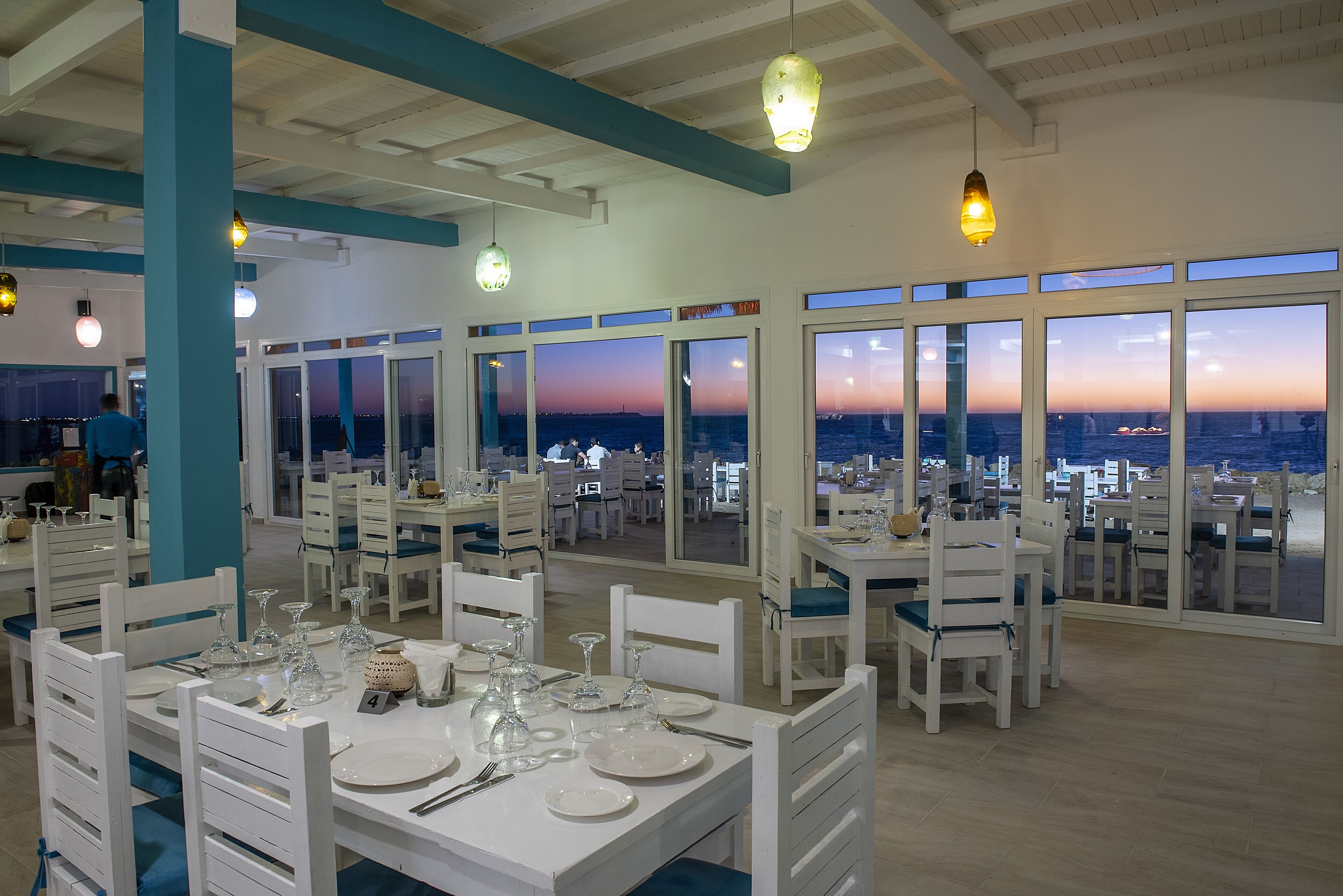 A restaurant with white tables and chairs, large windows with a city view, and hanging pendant lights.