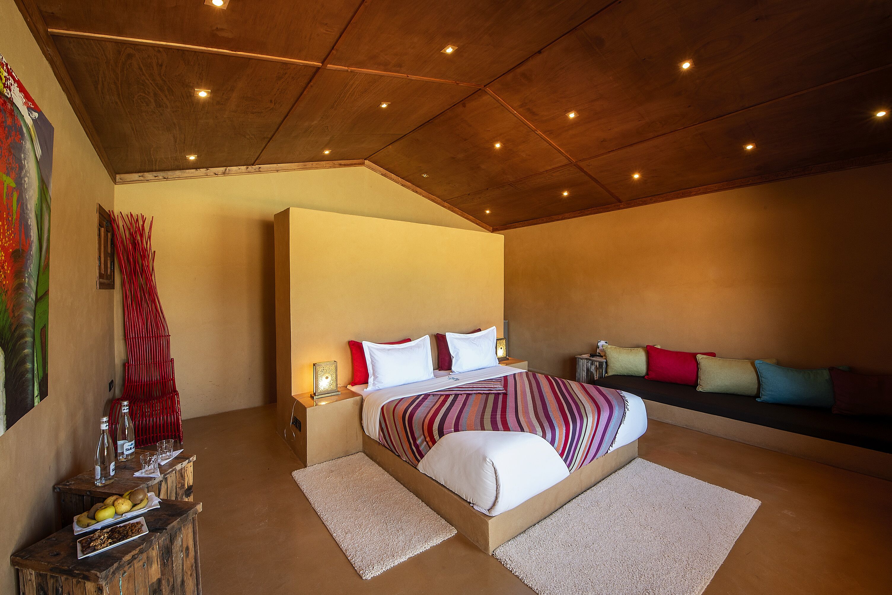 Un dormitorio con una cama grande, mesitas de noche, un banco con cojines y un cuadro en la pared.
