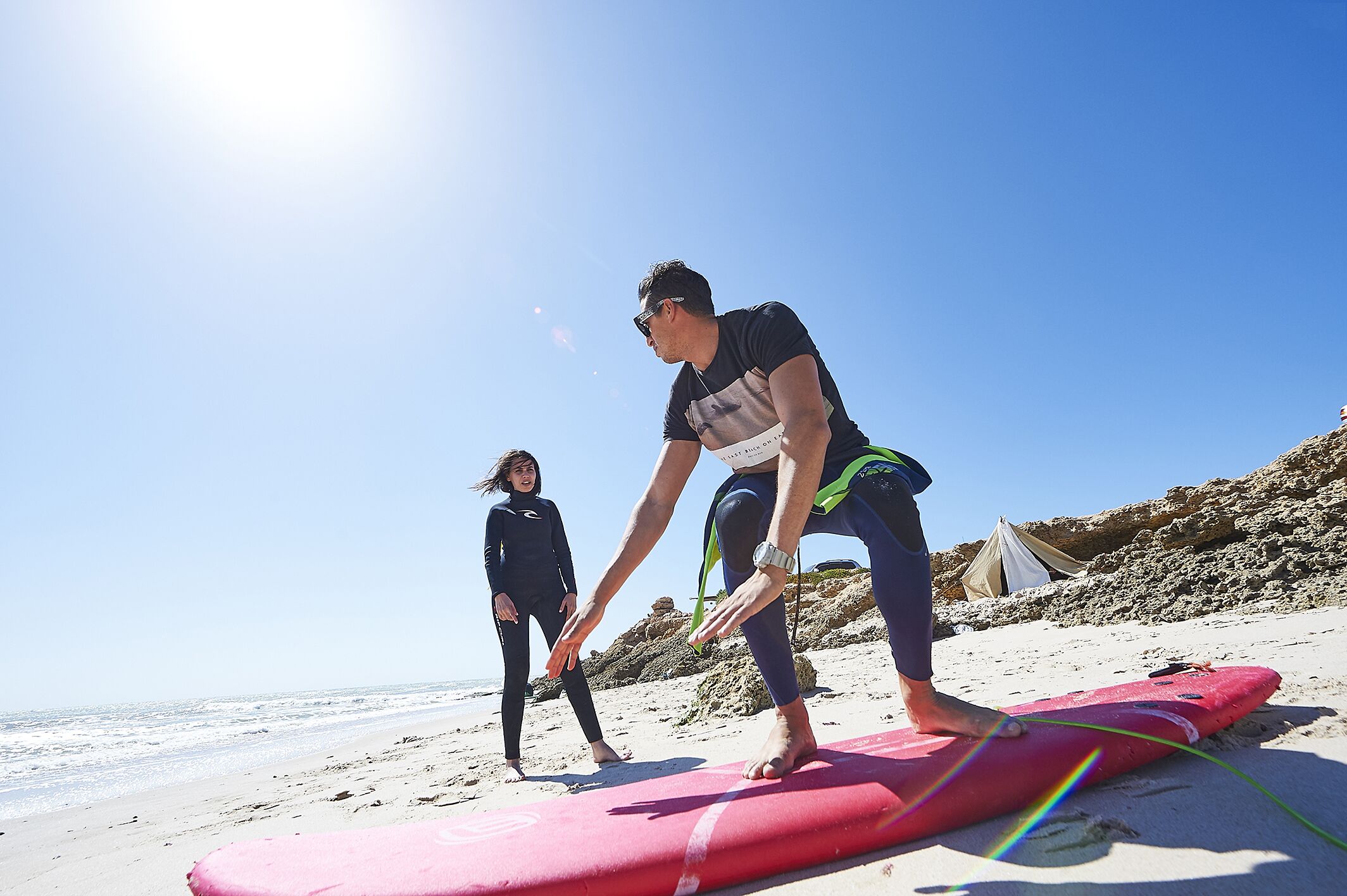 Una persona con traje de neopreno en una playa, con una tabla de surf y una tienda de campaña al fondo.