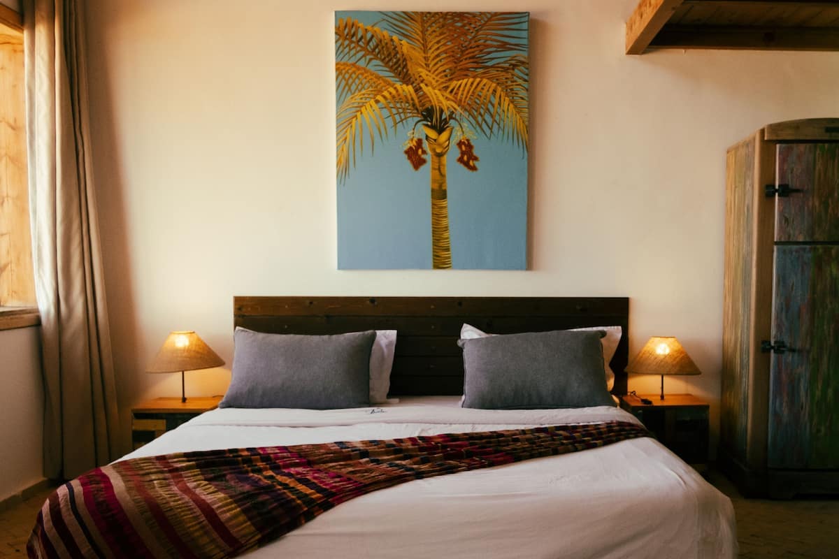 Une chambre à coucher avec un lit, deux oreillers, une couverture, une table de chevet avec une lampe, une armoire et un tableau représentant un palmier accroché au mur.
