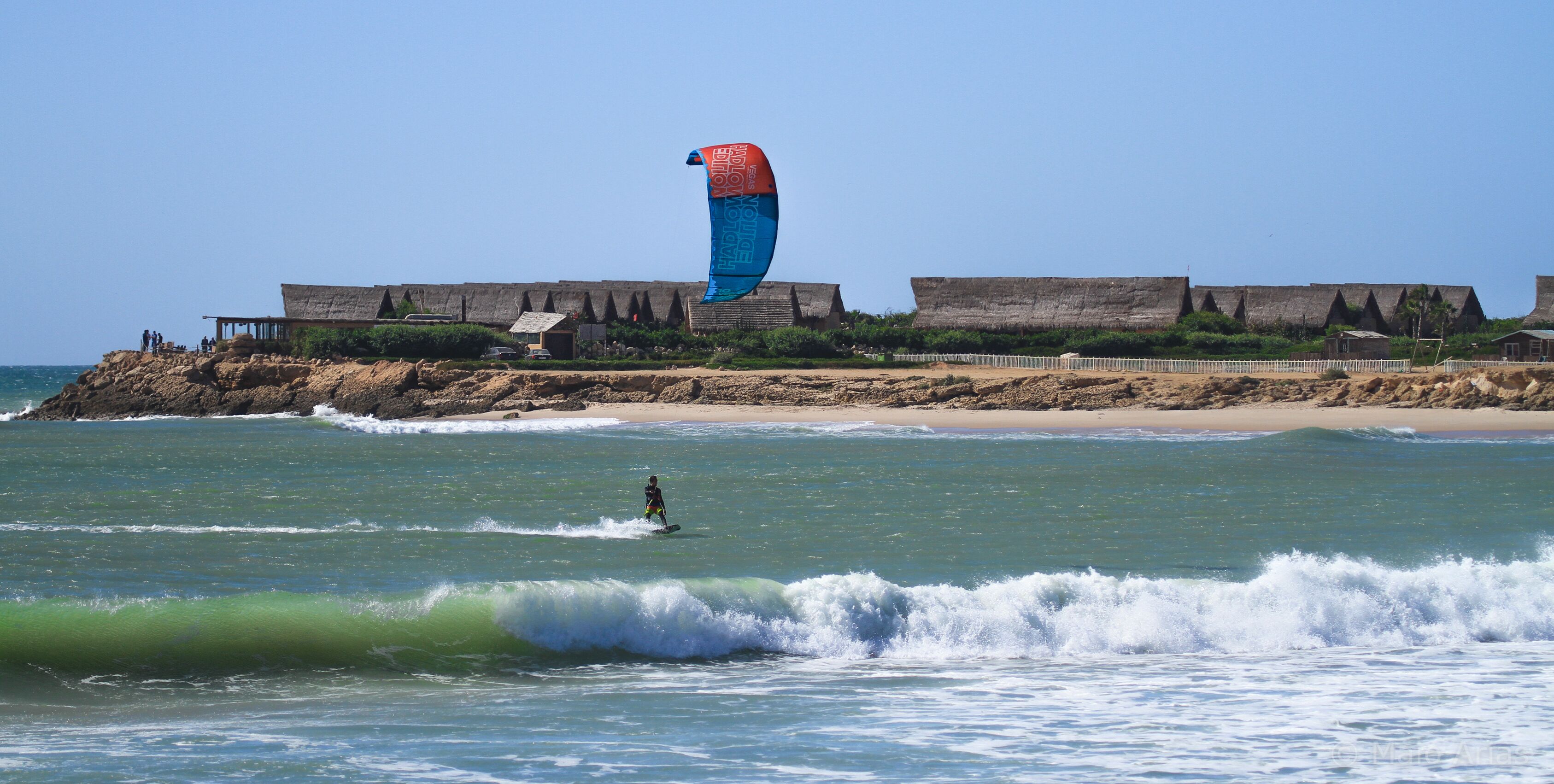 Ein Strand mit einer Person, die Windsurfen geht, einer großen, blau-roten Drachen und einem Gebäude mit einem Strohdach im Hintergrund.