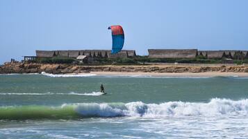 Ein Strand mit einer Person, die Windsurfen geht, einer großen, blau-roten Drachen und einem Gebäude mit einem Strohdach im Hintergrund.
