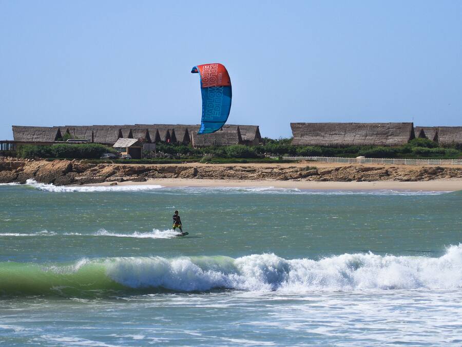 Ein Strand mit einer Person, die Windsurfen geht, einer großen, blau-roten Drachen und einem Gebäude mit einem Strohdach im Hintergrund.