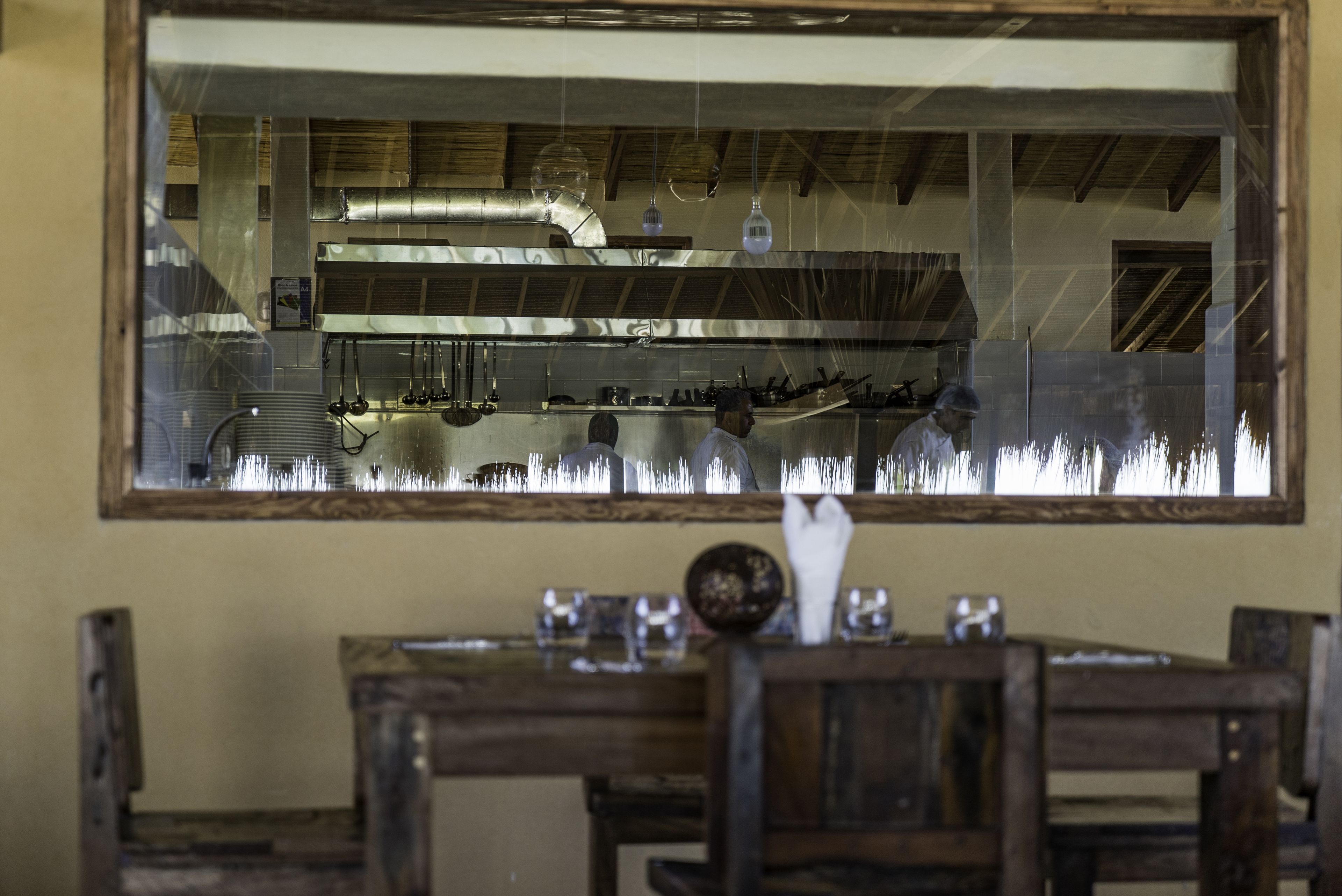 A dining area with a wooden table and chairs, a large mirror reflecting a kitchen with stainless steel appliances and hanging light fixtures.
