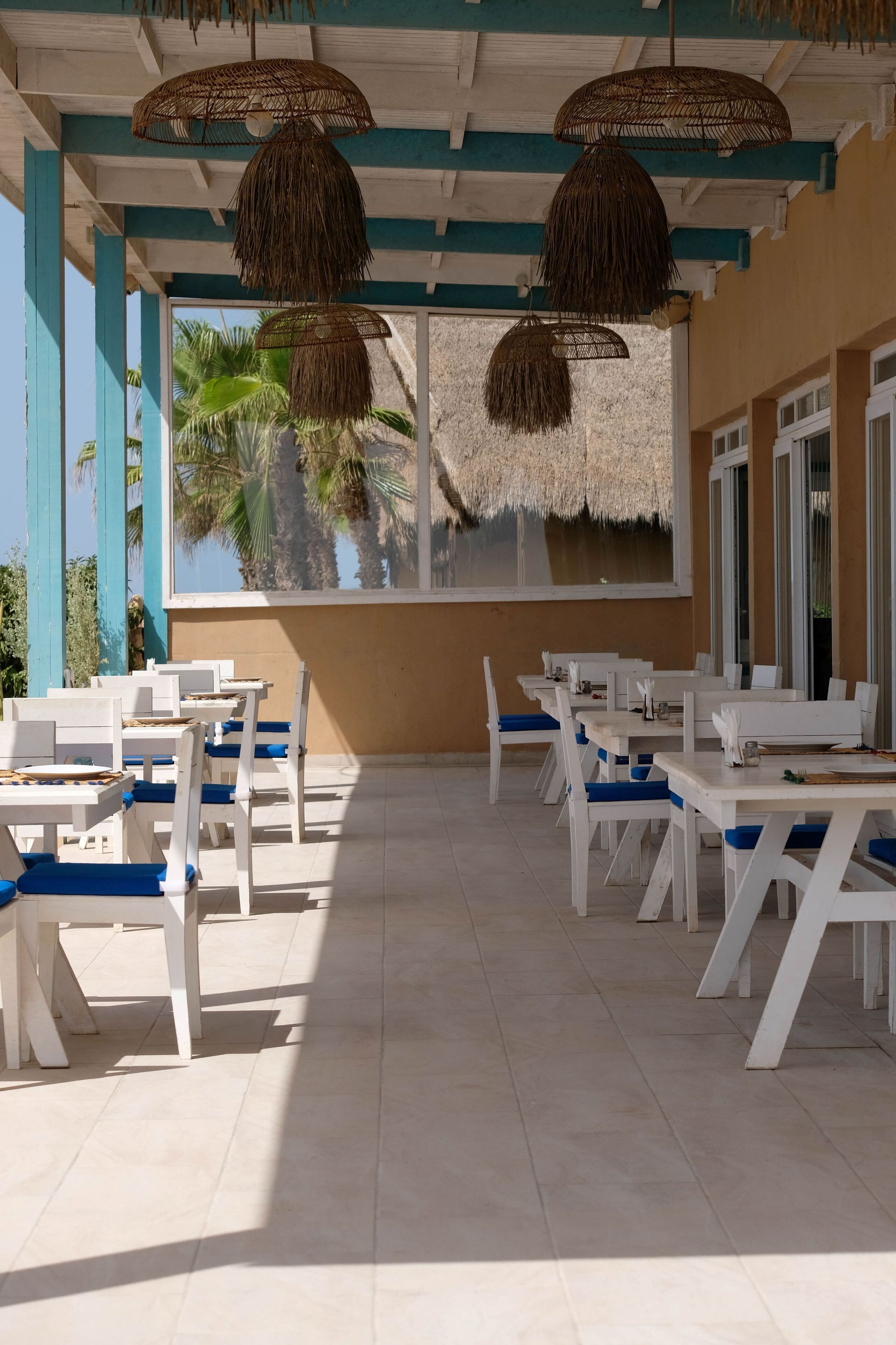 Un espace de restauration en plein air avec des tables blanches et des chaises bleues, des lampes suspendues en osier et une vue sur les palmiers à travers de grandes fenêtres.