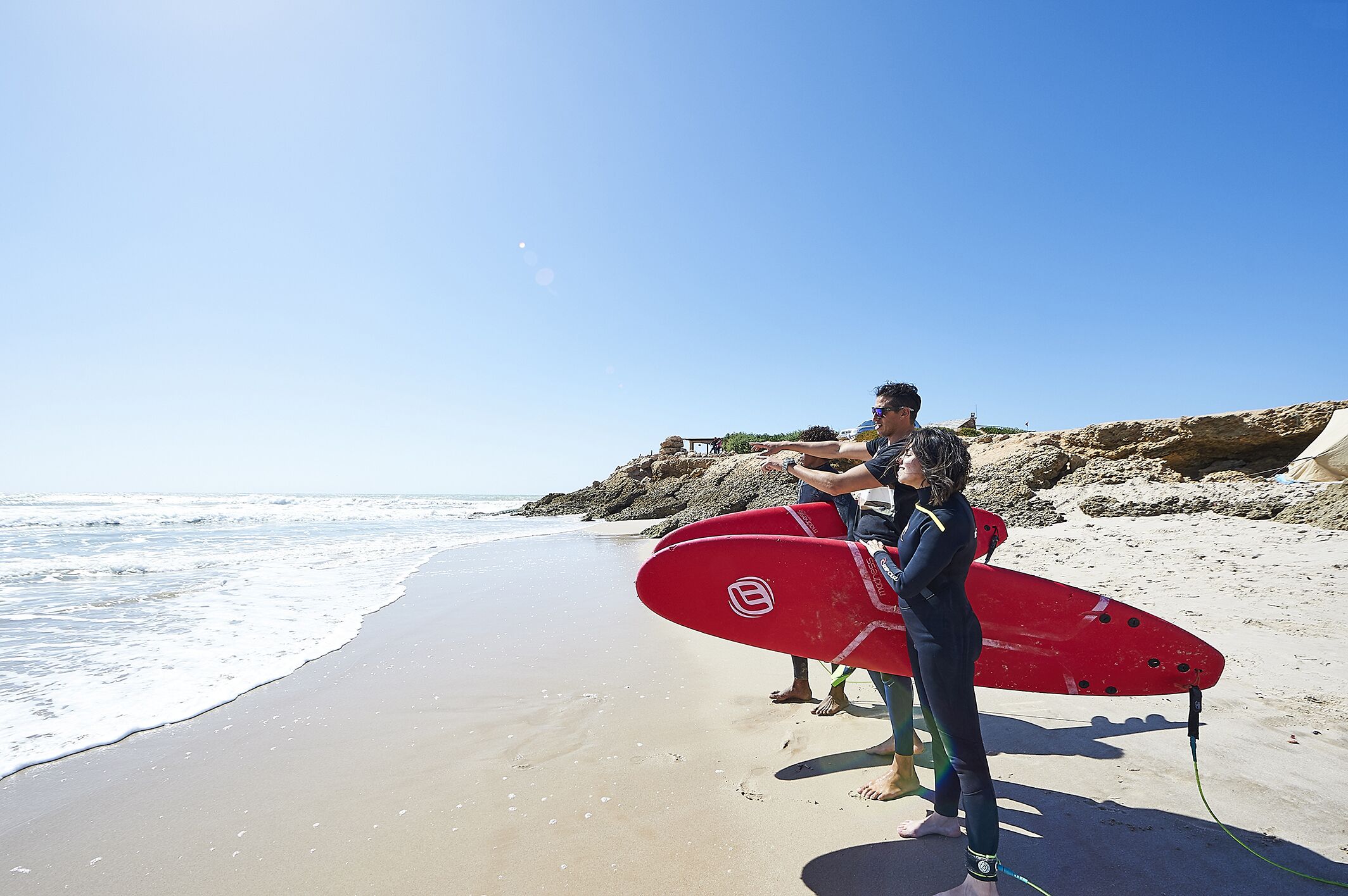 Una playa con cielo despejado, una tabla de surf roja y terreno rocoso.