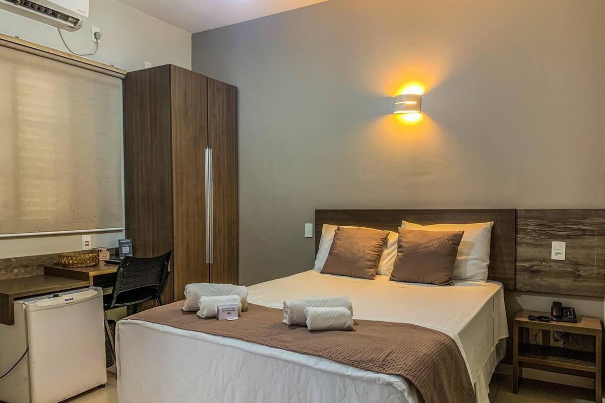 A hotel room with a bed, bedside table, desk, chair, and a small refrigerator.