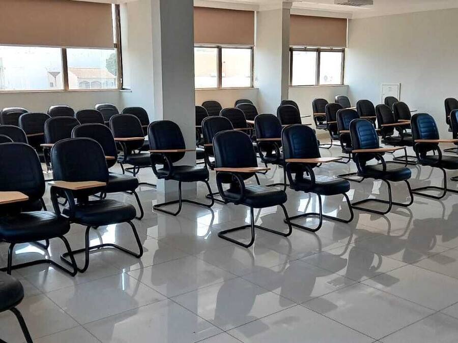 A classroom with rows of black chairs and desks, large windows, and fire extinguishers on the wall.