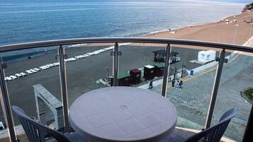A balcony with a round table and chairs, overlooking a beach with a promenade and beach huts.
