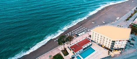 An aerial view of a coastal hotel with a swimming pool, beach chairs, and a red-roofed building.