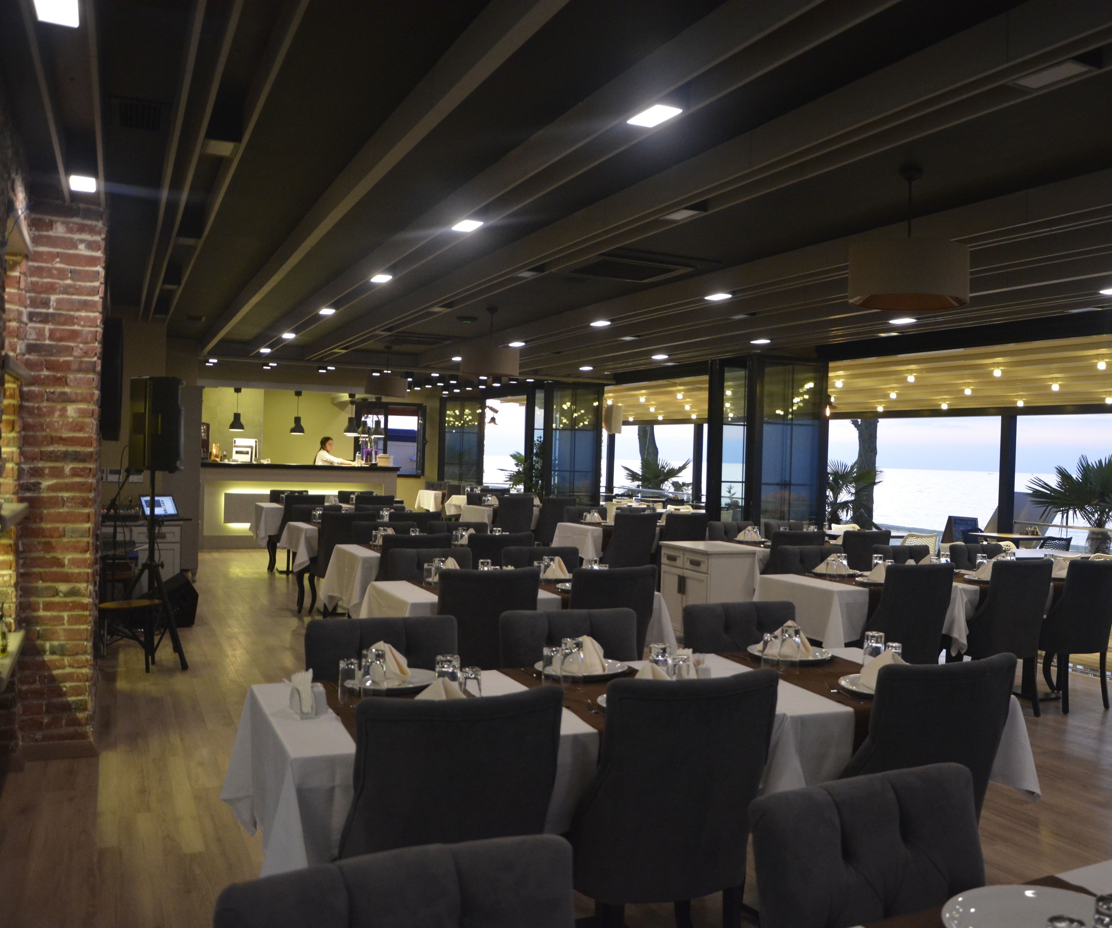 A restaurant with tables set for dining, a bar area, and large windows offering a view of the sea.