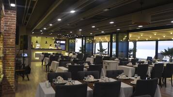 A restaurant with tables set for dining, a bar area, and large windows offering a view of the sea.