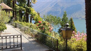 A pathway with a metal railing, surrounded by lush greenery and pink flowers, leading to a body of water with mountains in the background. - Barony Le Pergole (Lugano)