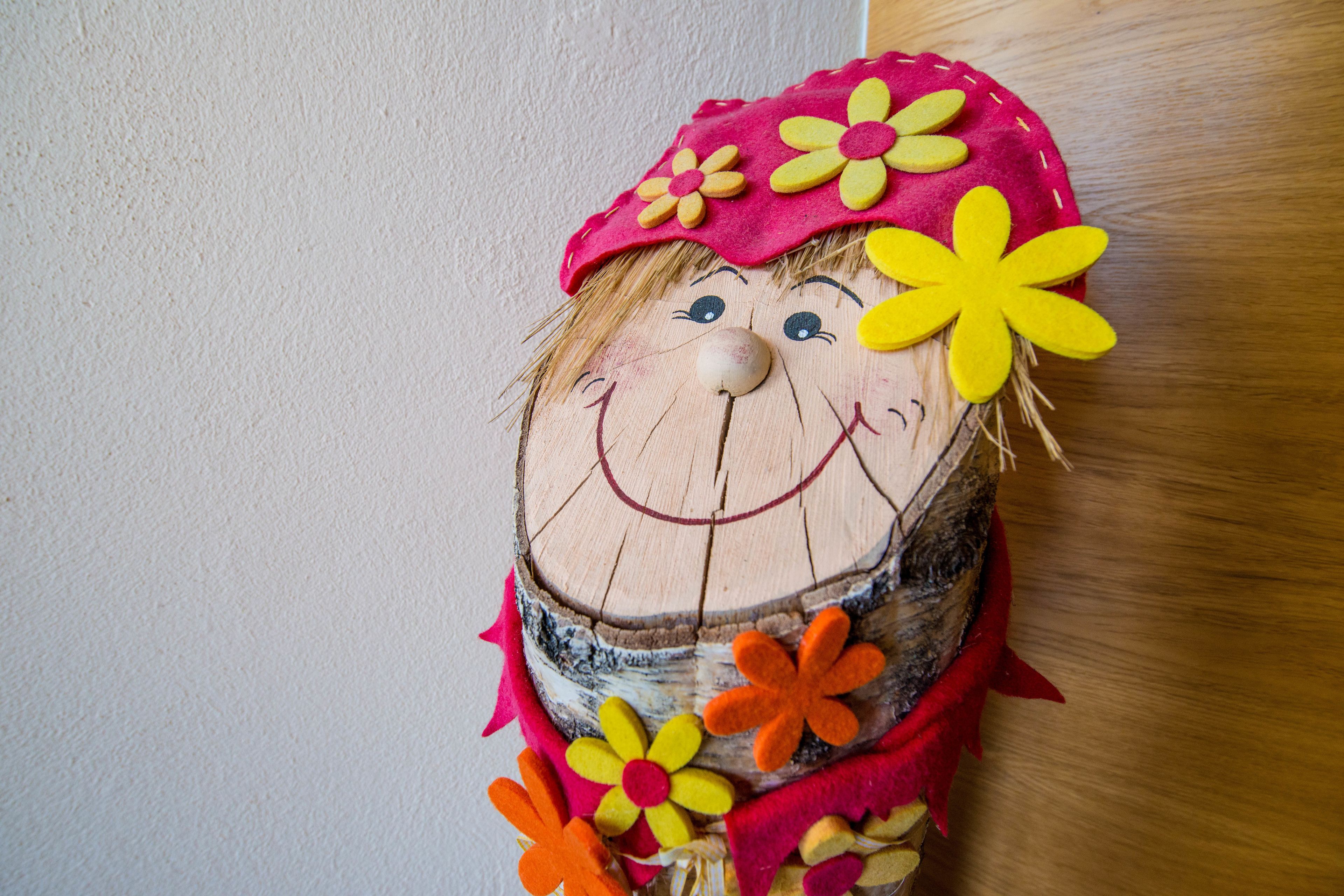 Eine rustikale Holzdeko mit einem lächelnden Gesicht, verziert mit Filzblumen und einem roten Hut.
