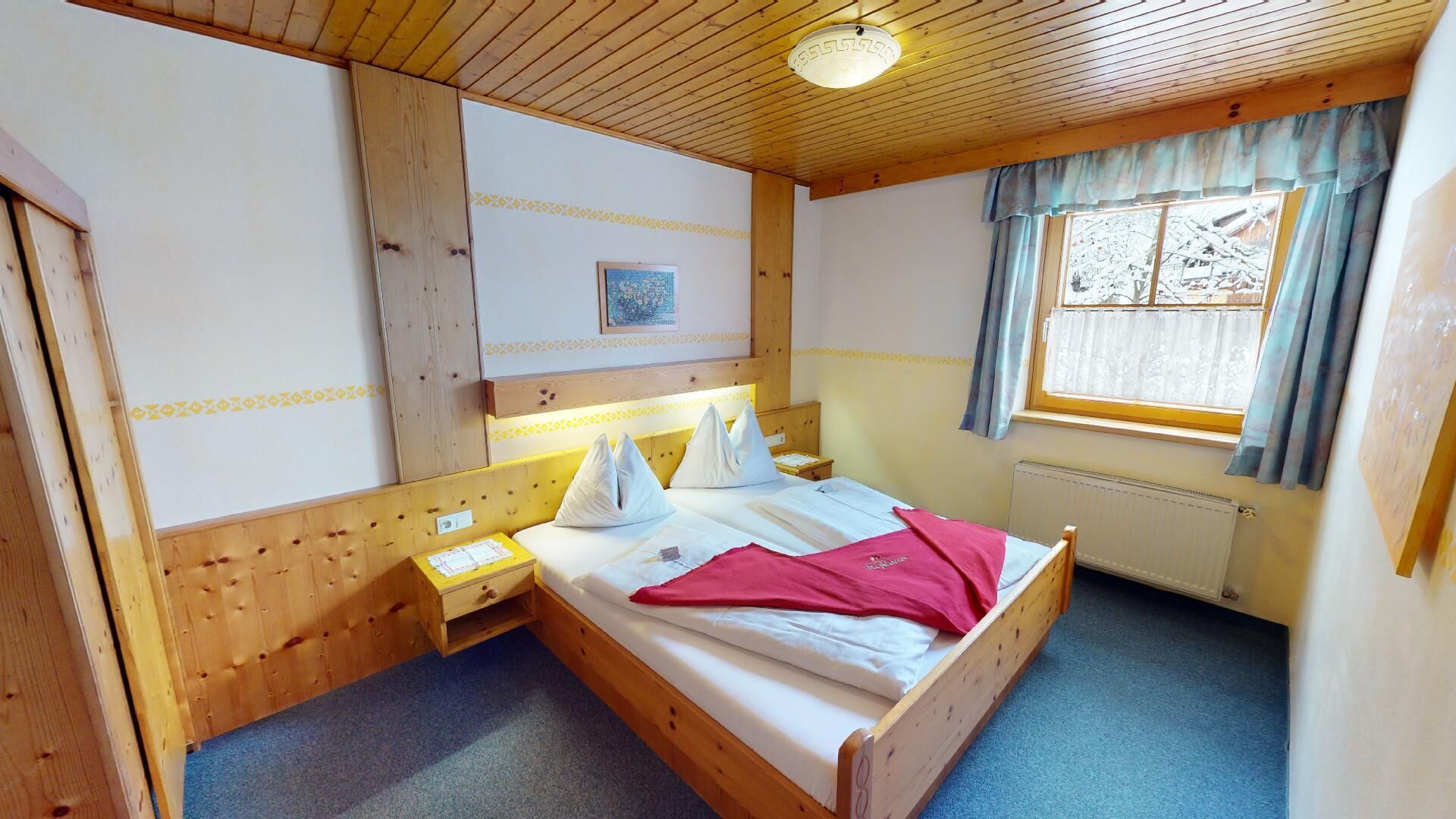 Habitación con paneles de madera, una cama, una mesita de noche, una ventana con cortinas y un radiador.
