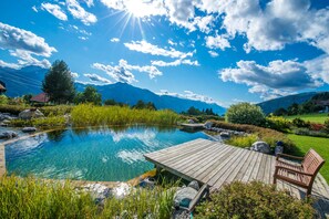 Una piscina cristalina con un muelle de madera y bancos, rodeada de exuberante vegetación y montañas al fondo.