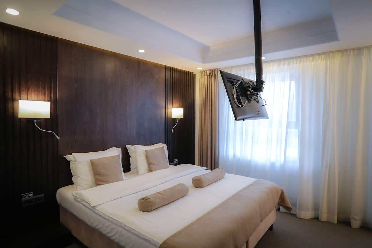 A hotel room with a large bed, two bedside lamps, a flat-screen TV mounted on the ceiling, and a window with sheer curtains.