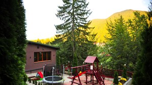 A playground with a swing set, slide, and climbing structure, surrounded by trees and a building with windows. - Royal Boutique Hotel Poiana Brasov (Brasov)