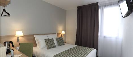 Habitación de hotel con cama, dos lámparas de noche, un televisor montado en la pared, un escritorio con una silla y una ventana con cortinas.