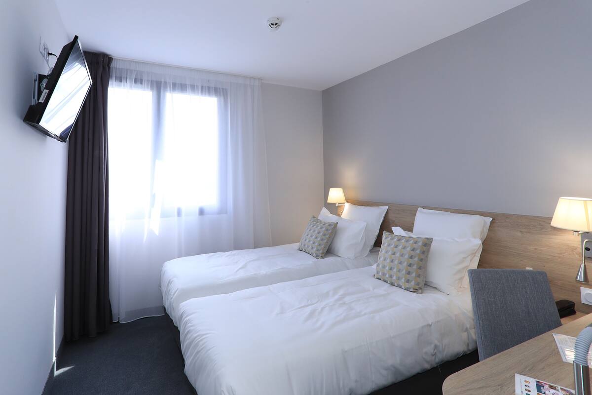 Une chambre d’hôtel avec deux lits, un bureau, une chaise, un téléviseur à écran plat fixé au mur et une fenêtre avec des rideaux.