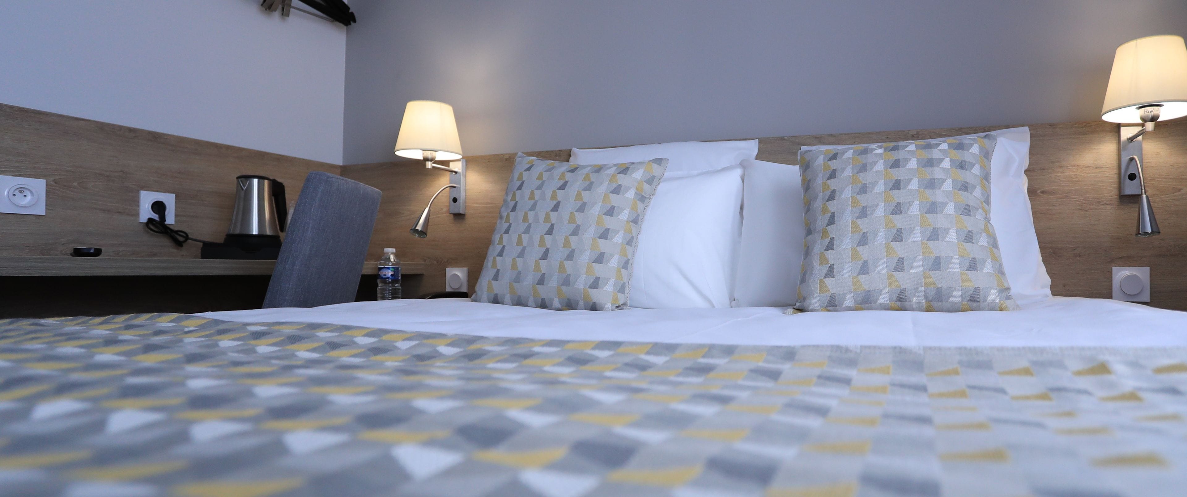 Habitación de hotel con una cama bien tendida, dos lámparas de noche, un cabecero de madera y una mesita con una botella de agua y una cafetera.