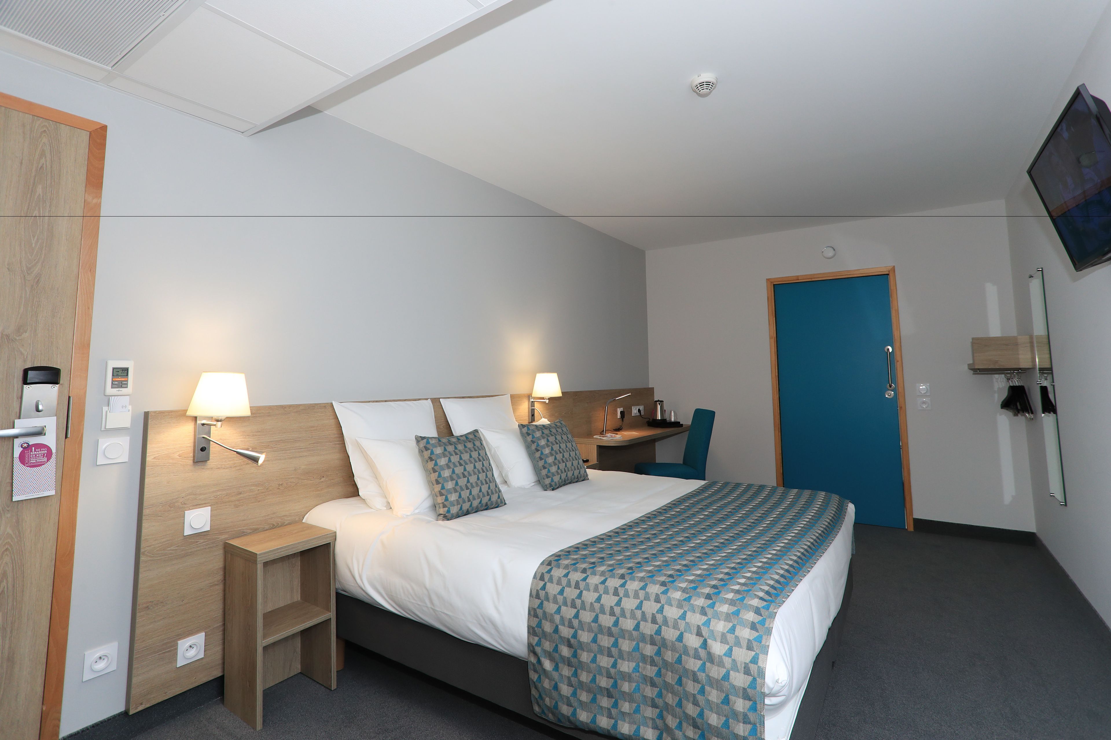 A hotel room with a bed, bedside tables, a desk, a chair, a TV, and a door.