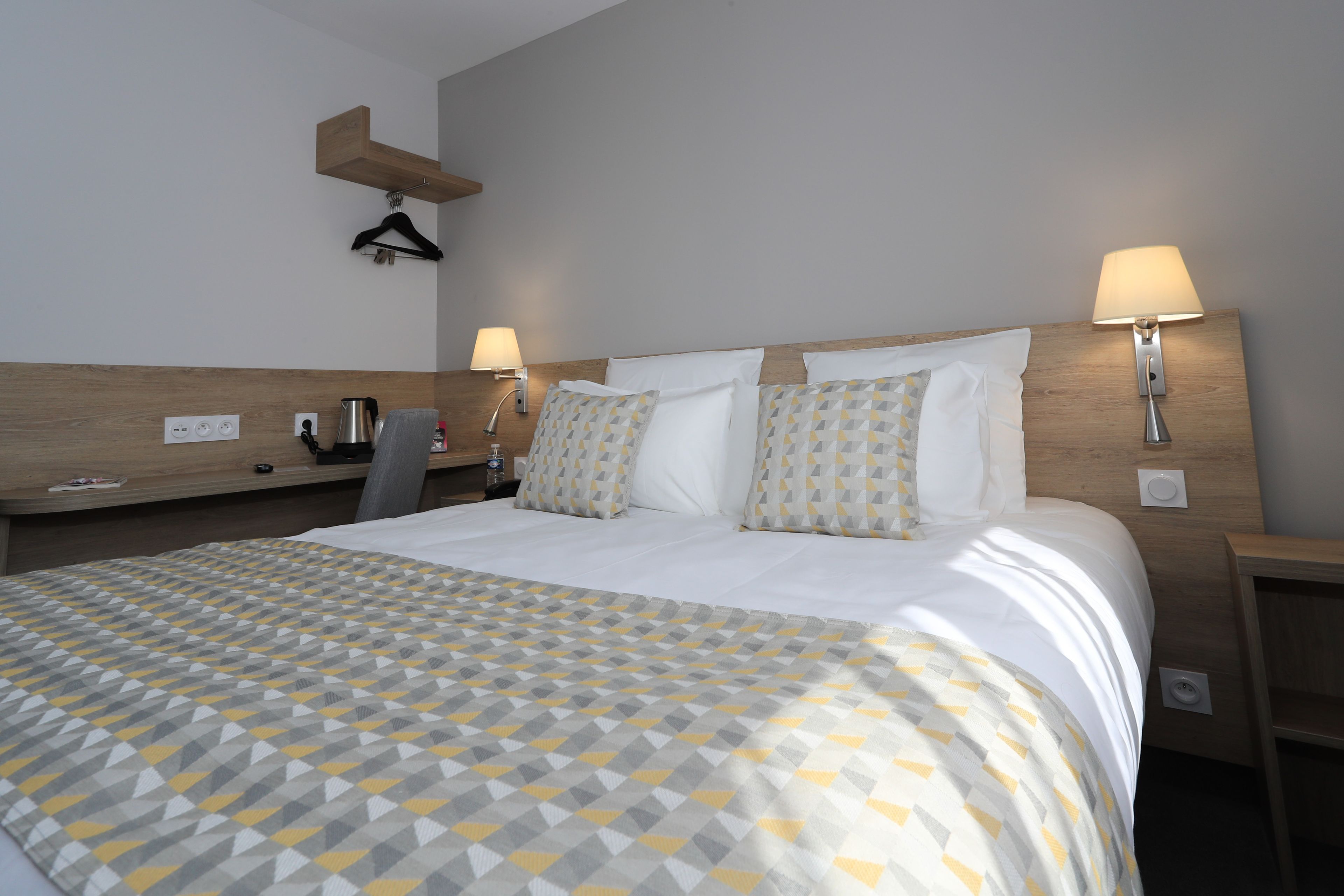 Ein Hotelzimmer mit einem ordentlich bezogenen Bett, Nachttischen mit Lampen, einem Holz-Kopfteil und einem an der Wand befestigten Regal.