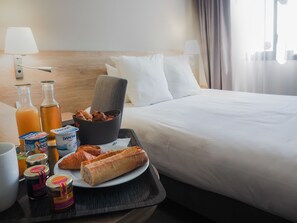 A hotel room with a neatly made bed, a tray with breakfast items including croissants, yogurt, and juice, and a bedside table with lamps and a bottle. - The Originals City, Hôtel de la Confluence (Damazan)