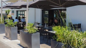 Un espace extérieur aménagé avec des tables et des chaises sous des parasols, entouré de jardinières remplies de plantes vertes.