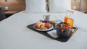 Un lit bien fait, avec du linge de lit blanc, un plateau contenant des aliments pour le petit-déjeuner, notamment des croissants, une bouteille de jus d’orange et une tasse.