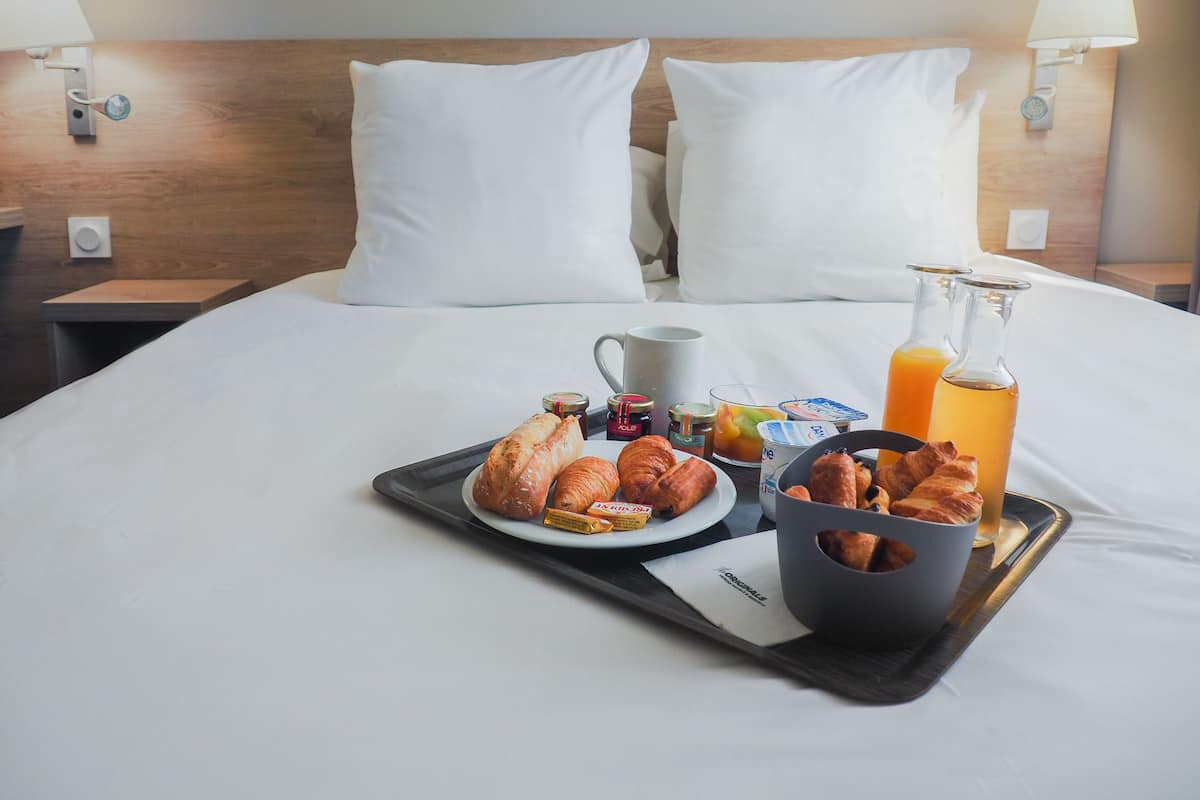 Un lit bien fait, avec du linge de lit blanc, un plateau contenant des aliments pour le petit-déjeuner, notamment des croissants, une bouteille de jus d’orange et une tasse.
