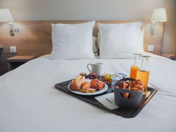 A neatly made bed with white linens, a tray with breakfast items including croissants, a bottle of orange juice, and a mug.