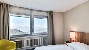 A hotel room with a bed, a chair, a nightstand, a lamp, and a window with a mountain view. - Hotel Pilatus Kulm (Kriens)