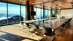 A conference room with a long table, chairs, a projector screen, and a large window offering a mountain view. - Hotel Pilatus Kulm (Kriens)