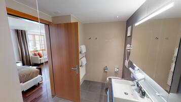 A modern bathroom with a large mirror, a sink, and a shower. There is a door leading to a bedroom with a bed and a window.