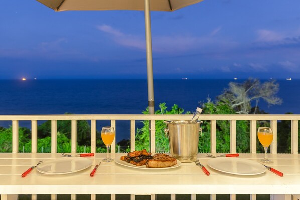 A dining table with plates, cutlery, and a pitcher of orange juice, overlooking a scenic ocean view.