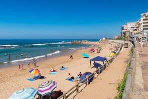 A beach with people sunbathing and swimming, a wooden fence, and buildings in the background.