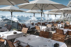 An outdoor dining area with tables and chairs, umbrellas, and a view of snow-capped mountains. - Hotel Pilatus-kulm (Kriens)