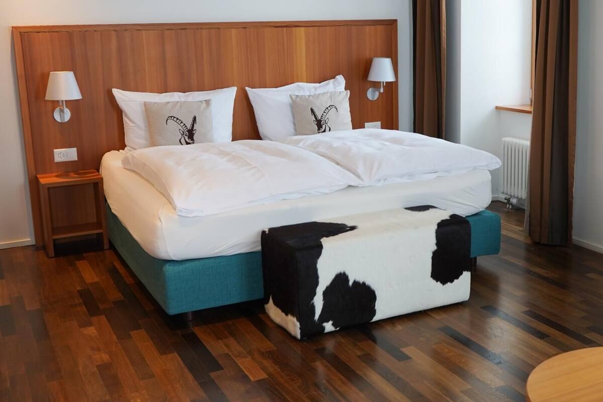 A hotel room with a wooden headboard, a bed with white bedding, two nightstands with lamps, a cow-patterned ottoman, and a wooden floor.