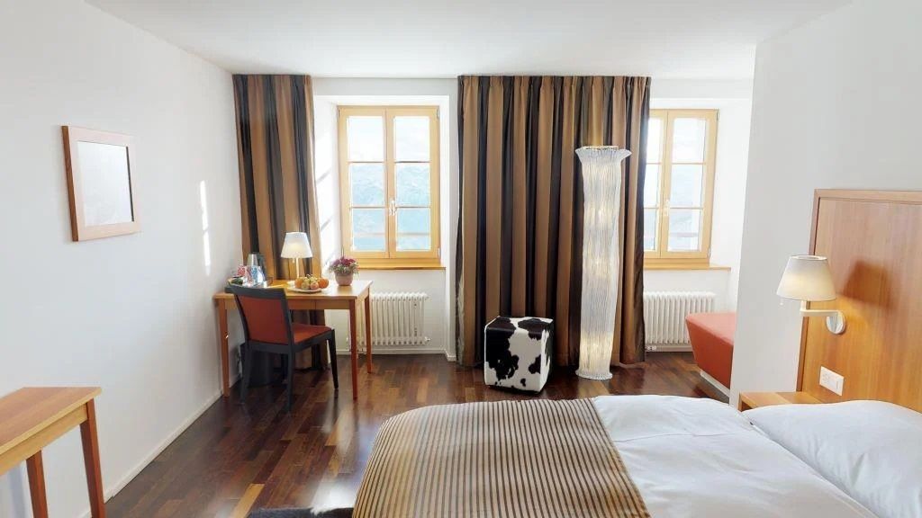 A hotel room with a bed, a desk, a chair, a lamp, a radiator, and a window with curtains.