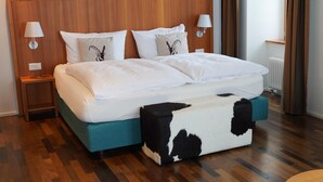 Ein Hotelzimmer mit einem Holzkopfteil, einem Bett mit weißer Bettwäsche, zwei Nachttischen mit Lampen, einem Zebra-Muster-Hocker und einem Holzboden.