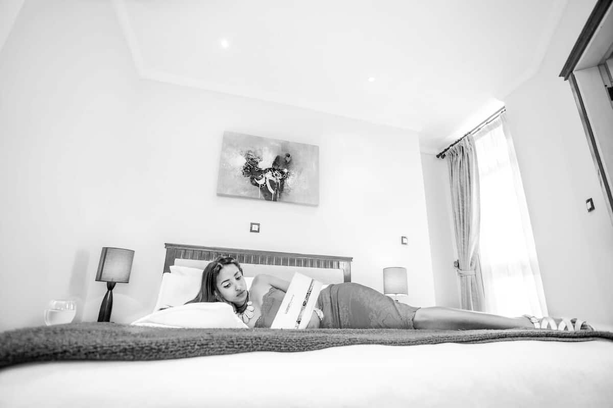A hotel room with a bed, two bedside lamps, a ceiling light, a painting on the wall, and a window with curtains.