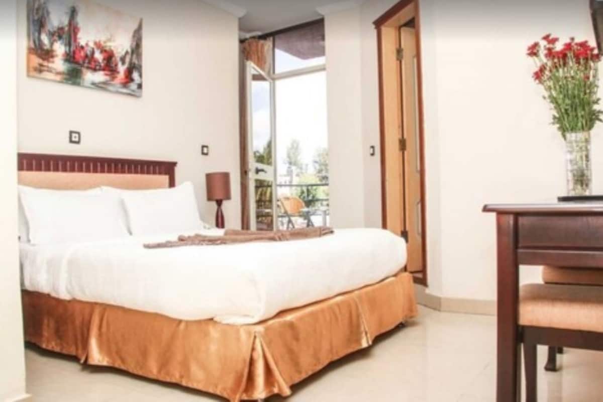 A bedroom with a bed, a desk, and a chair. There is a window with a view of the outdoors.